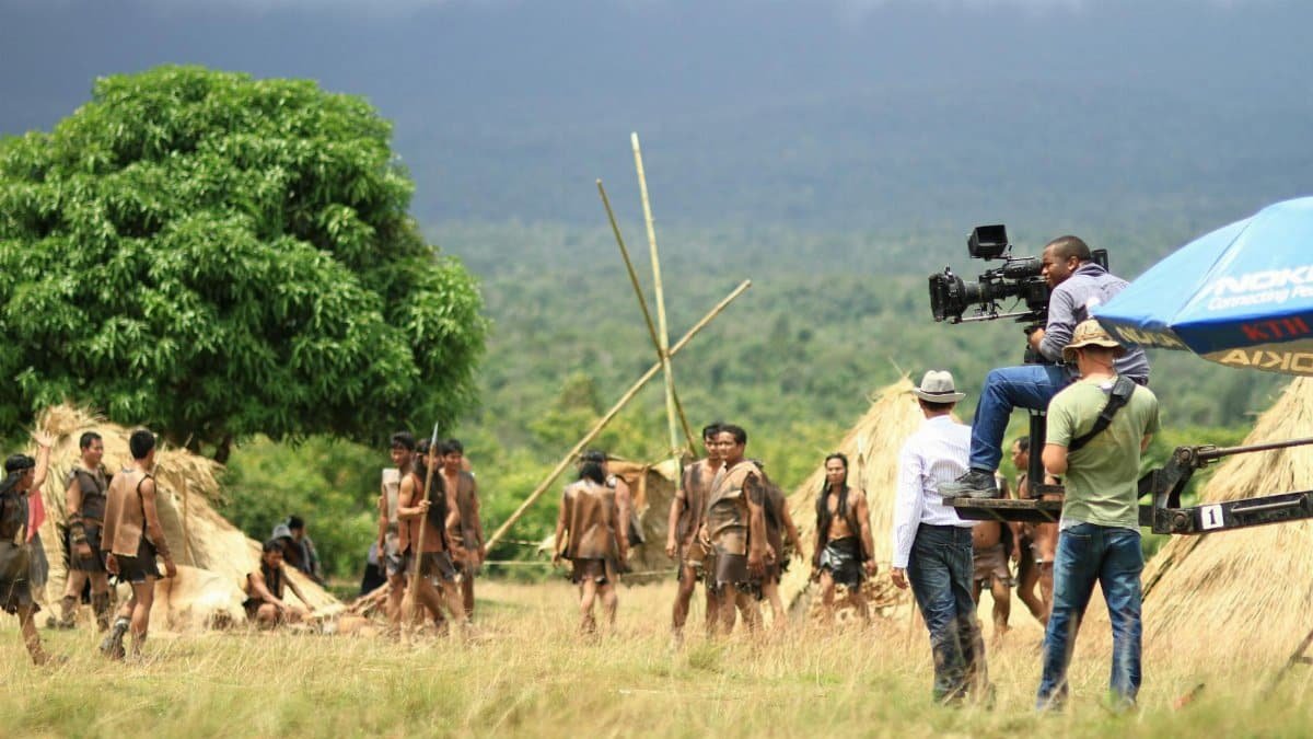 Behind the scenes view of a film crew capturing a scene on an outdoor set with actors and natural scenery.