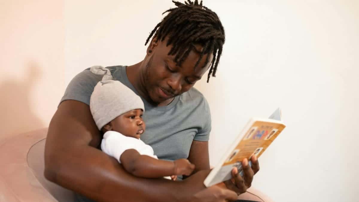 A father with dreadlocks reads a book to his baby, creating a bonding moment.