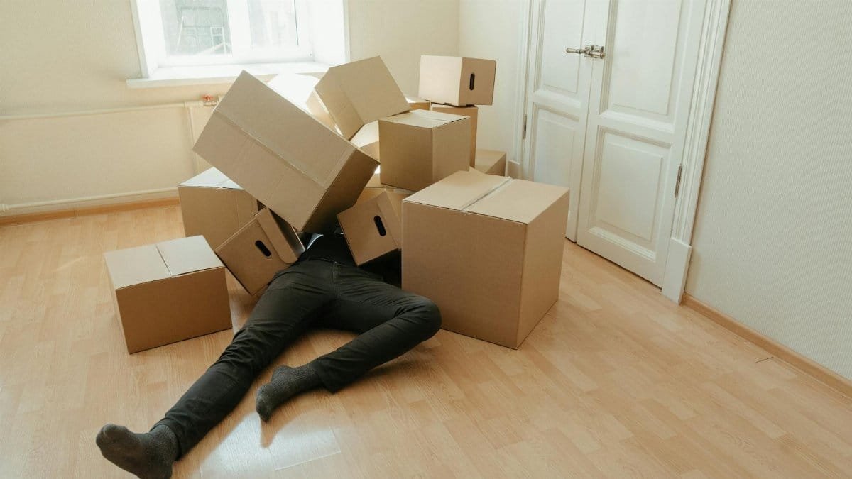 A person collapsed under stacks of cardboard boxes in an empty room, illustrating the stress of moving.