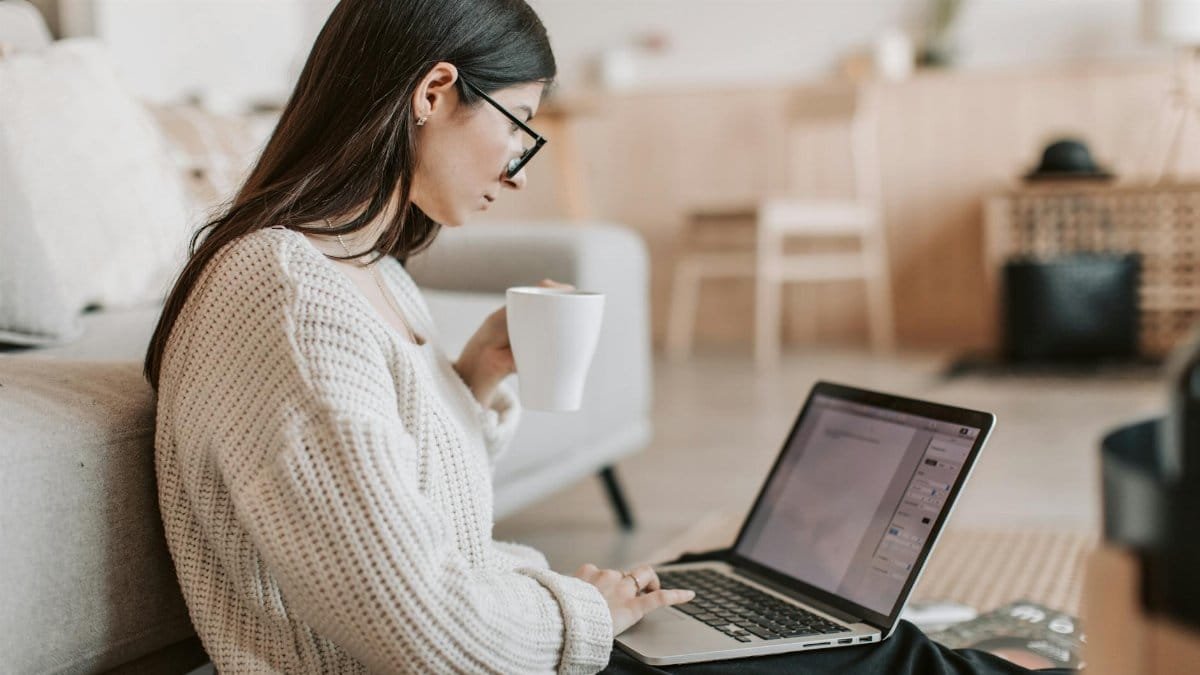 Side view of female freelancer in warm sweater and eyeglasses drinking tea from white ceramic cup while sitting on floor near sofa with netbook on legs while creating document for remote work project