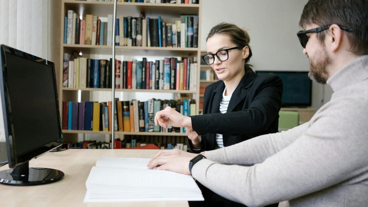 A woman helps a blind man read Braille at a desk in a library setting.