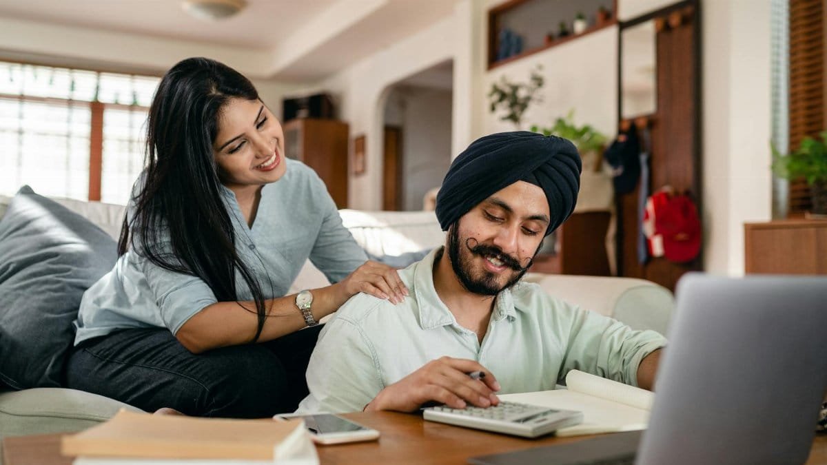 Positive ethnic man sitting at coffee table with laptop and smartphone and books and notebook and calculating while smiling ethnic wife sitting on sofa and supporting and watching behind