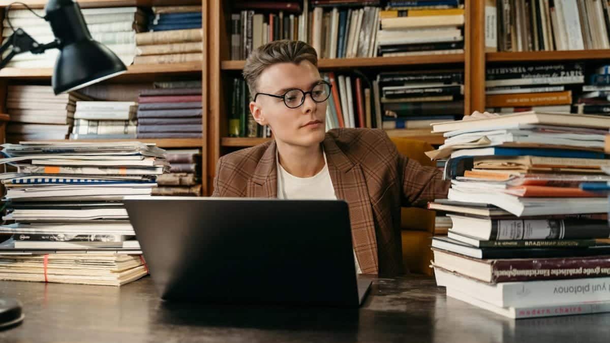 A young man with glasses working on a laptop surrounded by books in a library.
