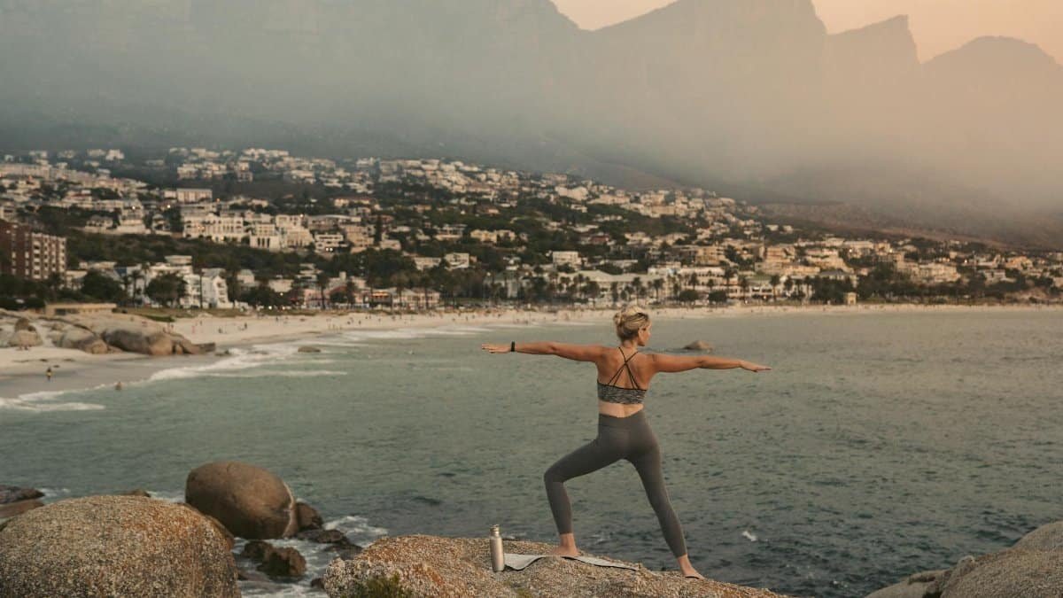 Woman practicing yoga warrior pose with scenic view of Cape Town coastline at sunset, promoting wellness.