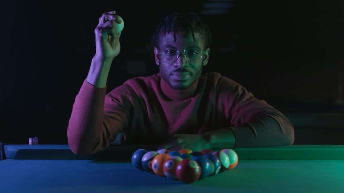 A man lines up a pool shot in a dimly lit room, holding a cue ball confidently.