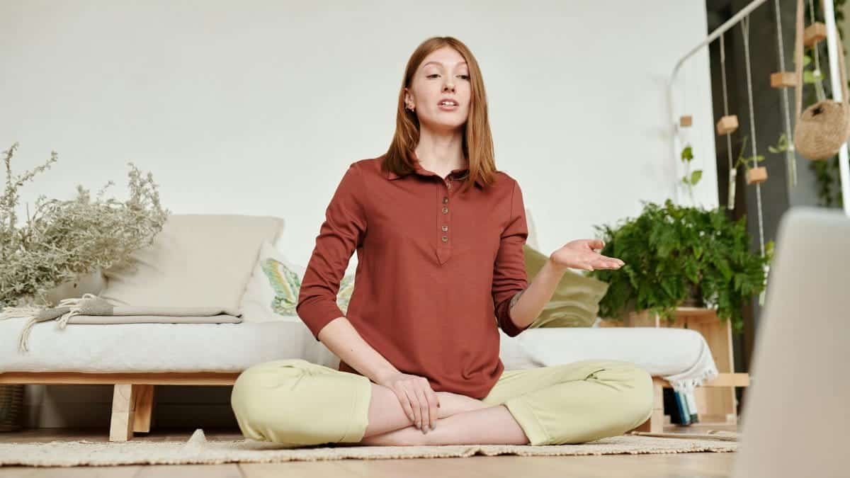 Woman practicing yoga in lotus position while attending an online class at home, surrounded by plants.