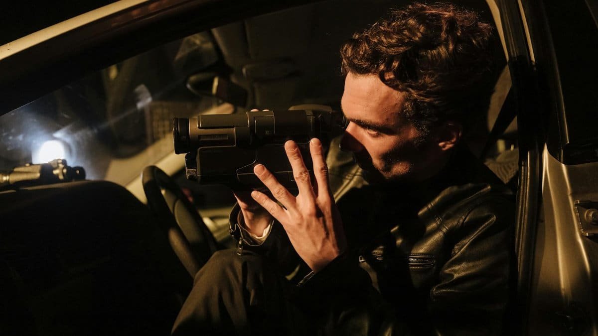 A man in a leather jacket uses a camcorder at night inside a car.