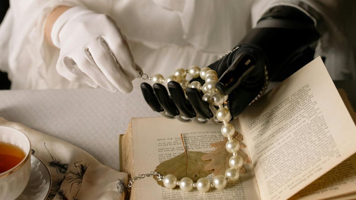 A graceful prosthetic hand holds a pearl necklace over an open book, blending technology and elegance.