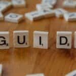 Scrabble tiles forming the word 'Guide' surrounded by scattered letters on a wooden table.
