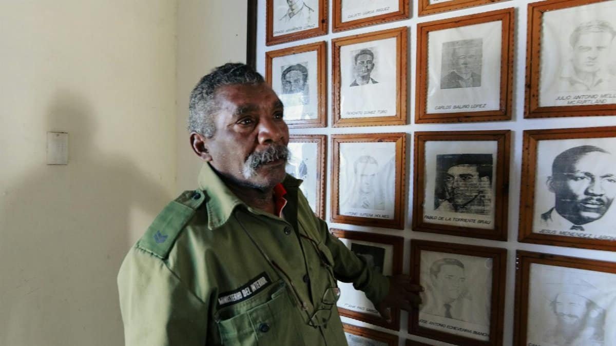An older military veteran in uniform, stands by a wall of historical black and white portraits indoors.