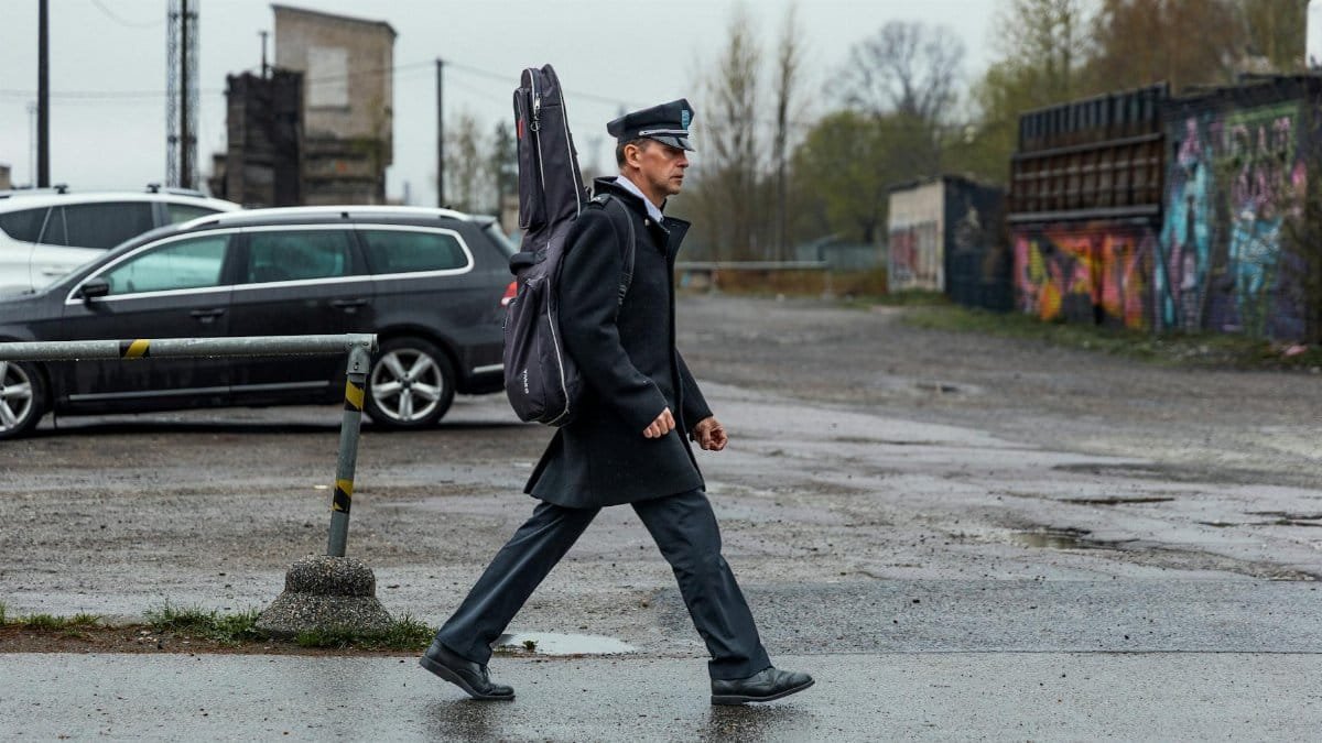 A man in uniform walks through an urban setting carrying a guitar case, showcasing street style.