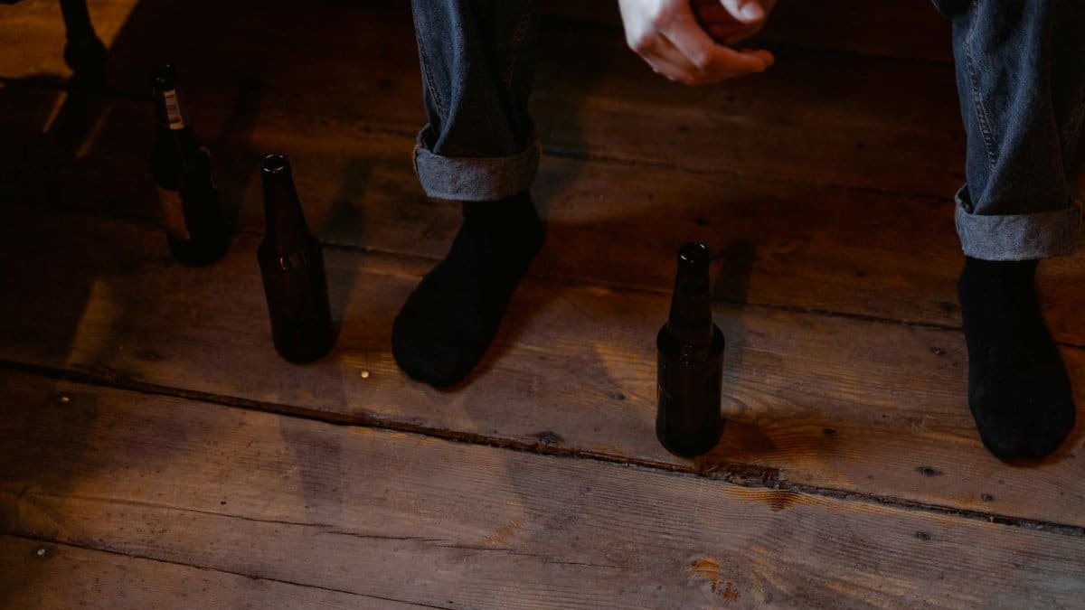 A person in denim and black socks sits on a wooden floor surrounded by beer bottles, reflecting a contemplative atmosphere.