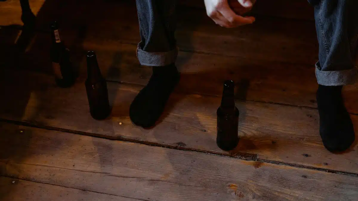 A person in denim and black socks sits on a wooden floor surrounded by beer bottles, reflecting a contemplative atmosphere.