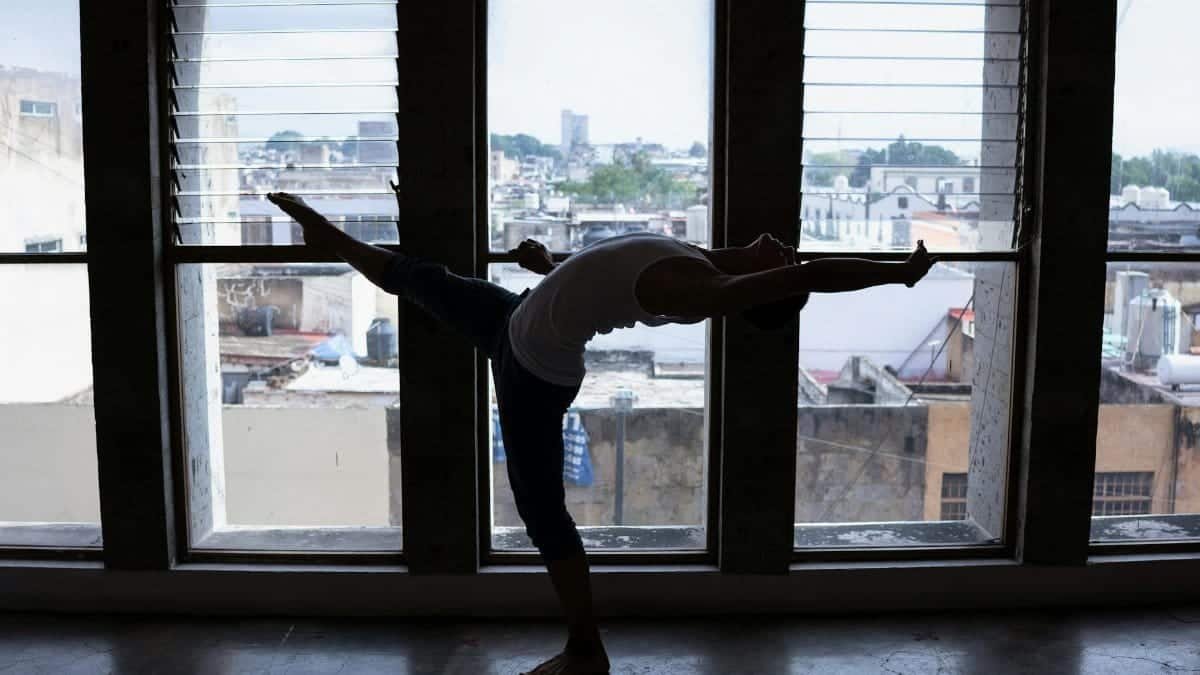 Backlit silhouette of a dancer performing a stretch in front of large urban windows, highlighting flexibility.