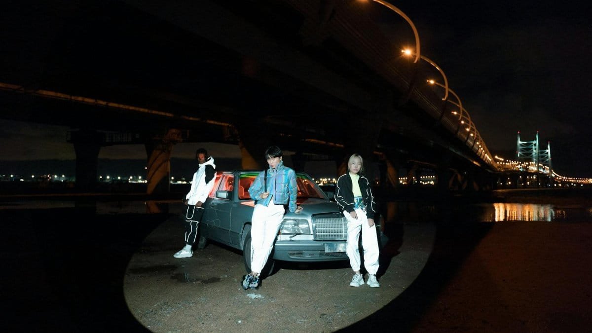 Three people standing in spotlight by a classic car under a city bridge at night.