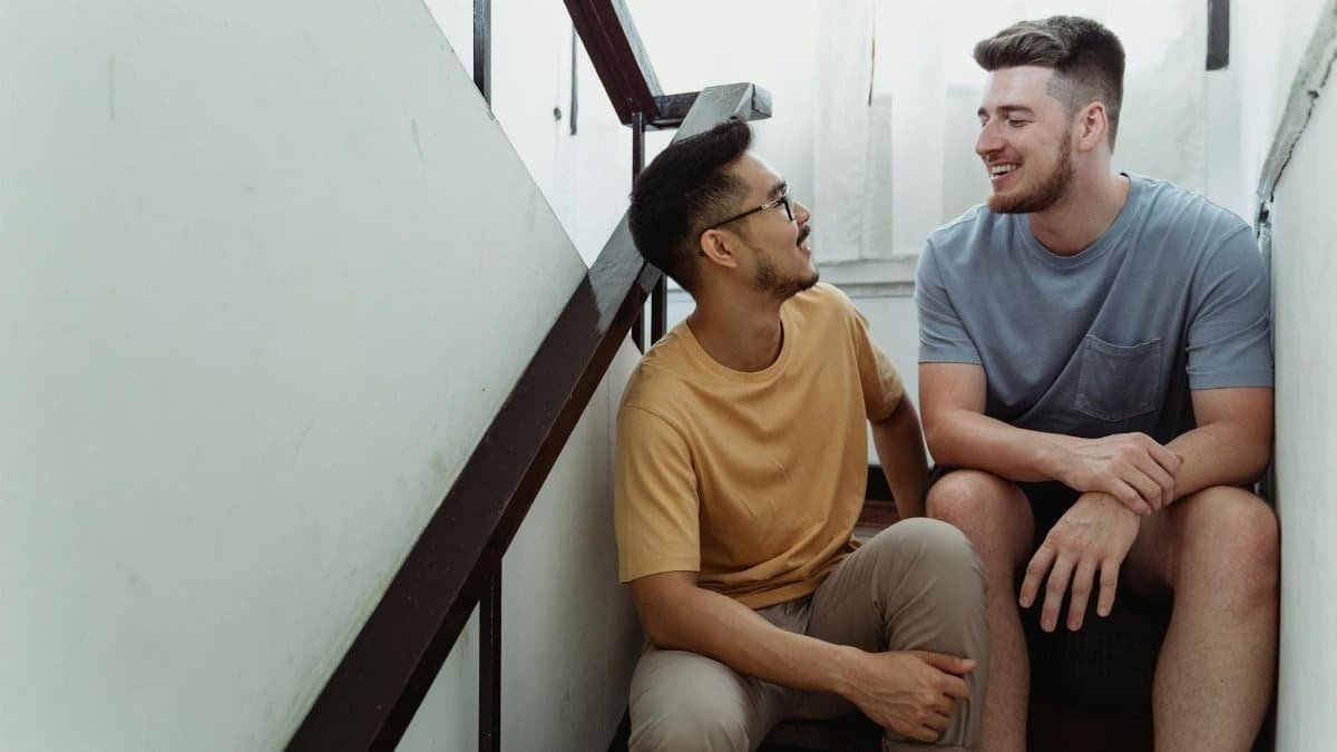 Loving LGBTQ couple smiling and sitting on wooden stairs indoors, enjoying a relaxed moment together.