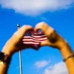 Hands form a heart shape around an American flag against a vibrant blue sky, symbolizing patriotism and freedom.