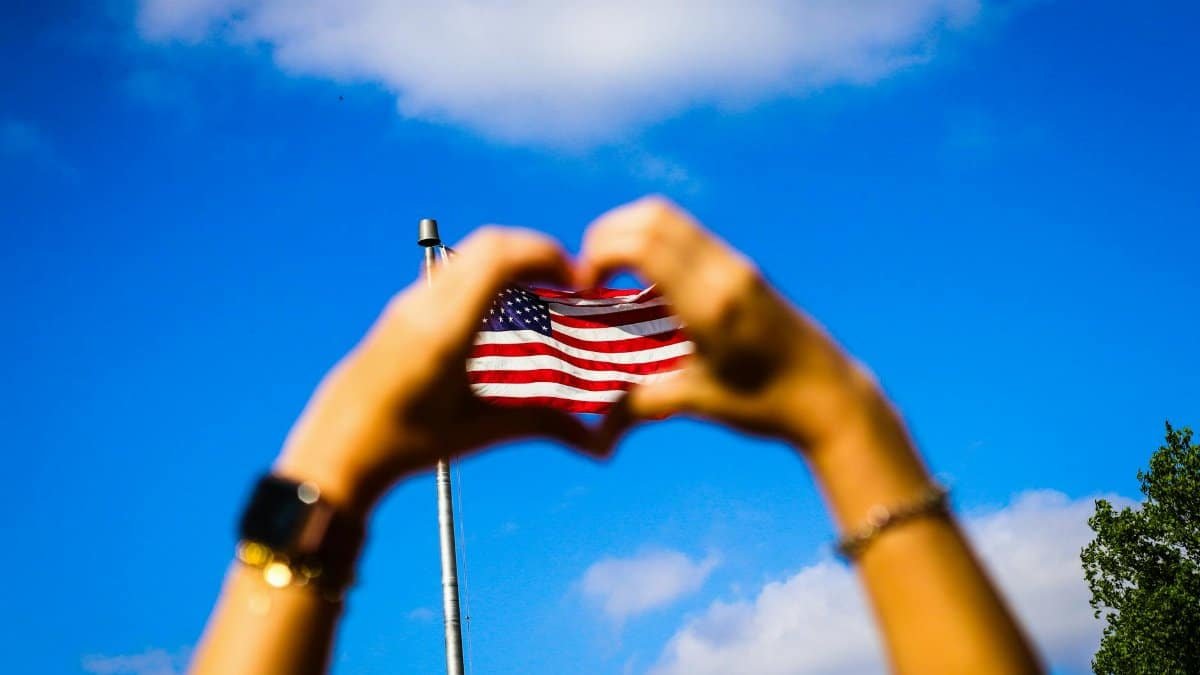 Hands form a heart shape around an American flag against a vibrant blue sky, symbolizing patriotism and freedom.