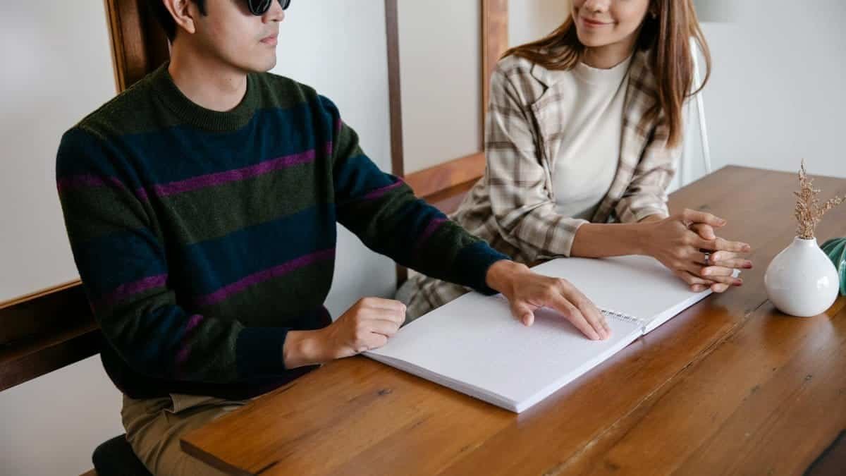 A visually impaired man reads a Braille book with a supportive woman sitting beside him indoors.