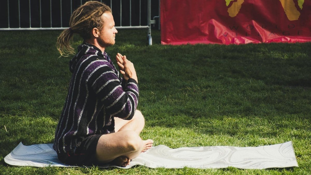 A man meditates in a seated yoga pose on a mat outdoors, enjoying the sunny day.