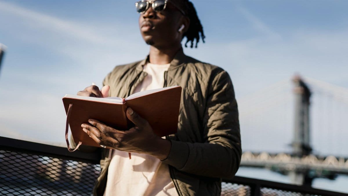 Man with dreadlocks writing in a notebook near a bridge, enjoying a calm outdoor setting.