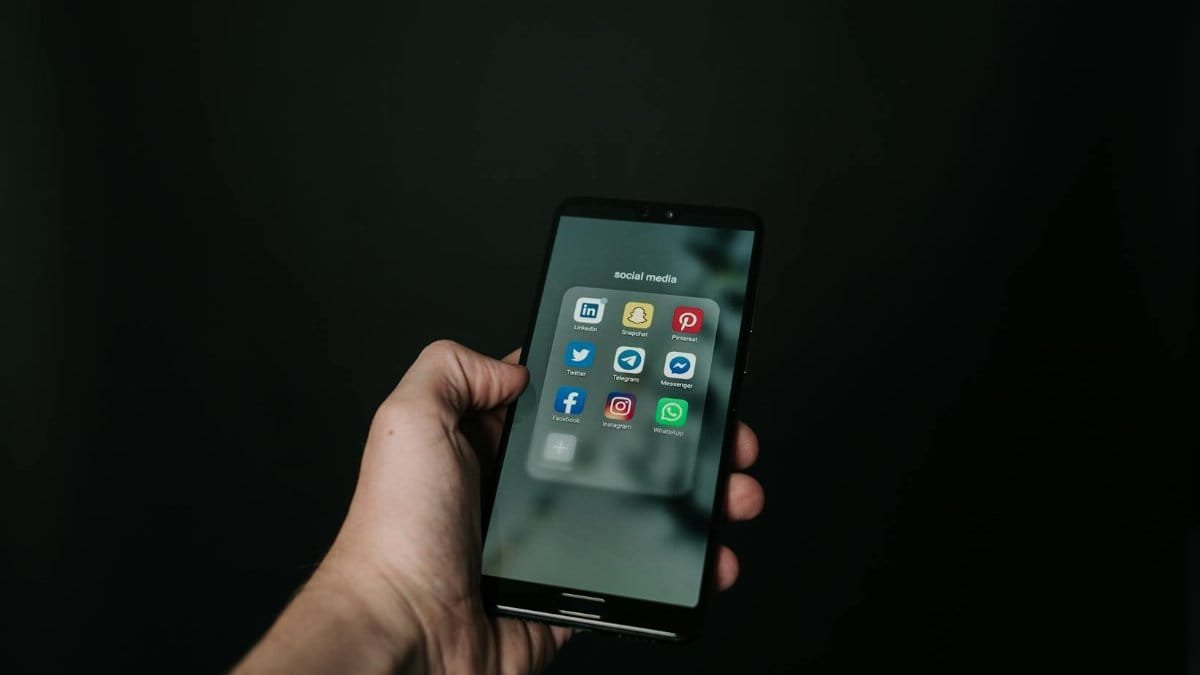 Close-up of a hand holding a smartphone displaying various social media app icons on a dark background.