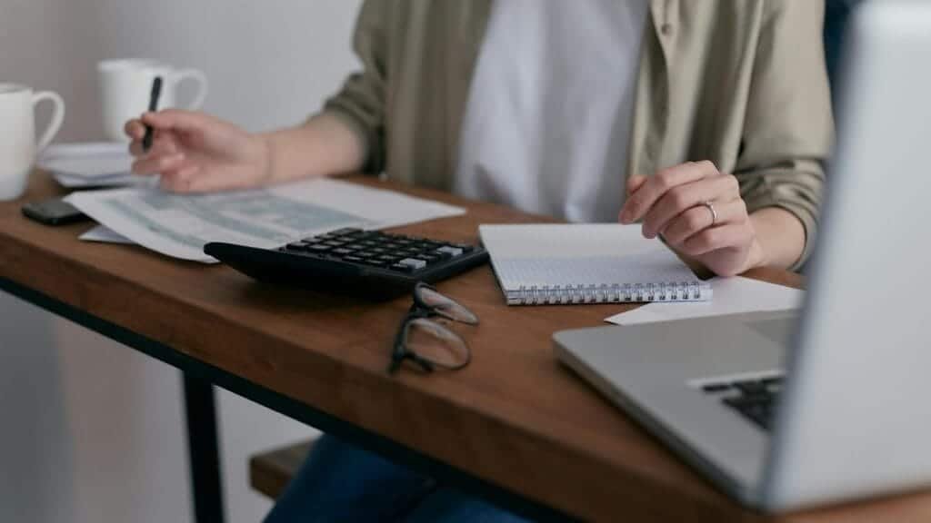 A woman manages finances at home, using a laptop and calculator on a wooden desk.