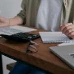 A woman manages finances at home, using a laptop and calculator on a wooden desk.