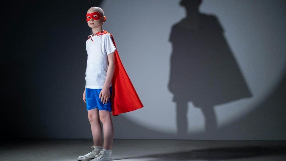 Child in a superhero costume poses confidently with a shadow on a studio wall, symbolizing strength and dreams.