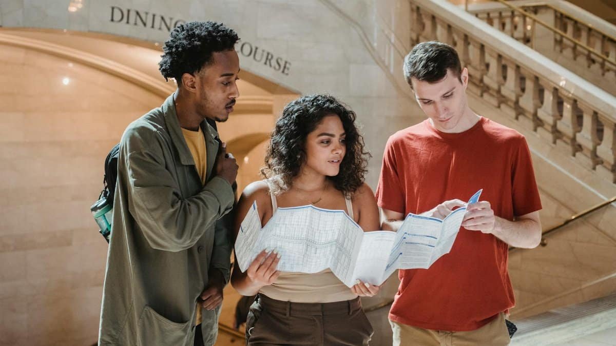 Focused young man pointing at map while searching for route with multiracial friends in Grand Central Terminal during trip in New York