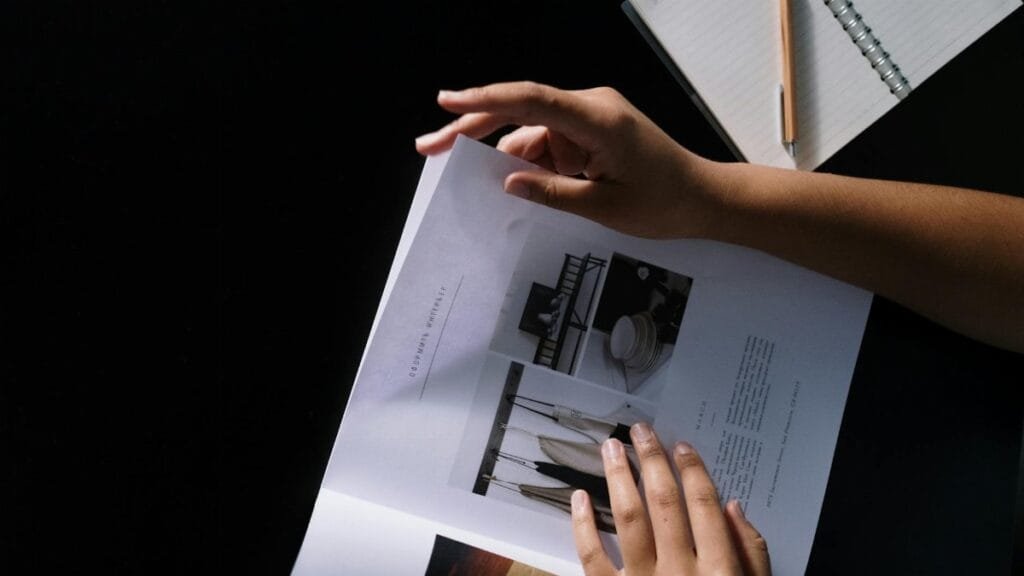 Hands flipping through a design magazine on a dark desk with a notebook and pencil.