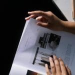 Hands flipping through a design magazine on a dark desk with a notebook and pencil.