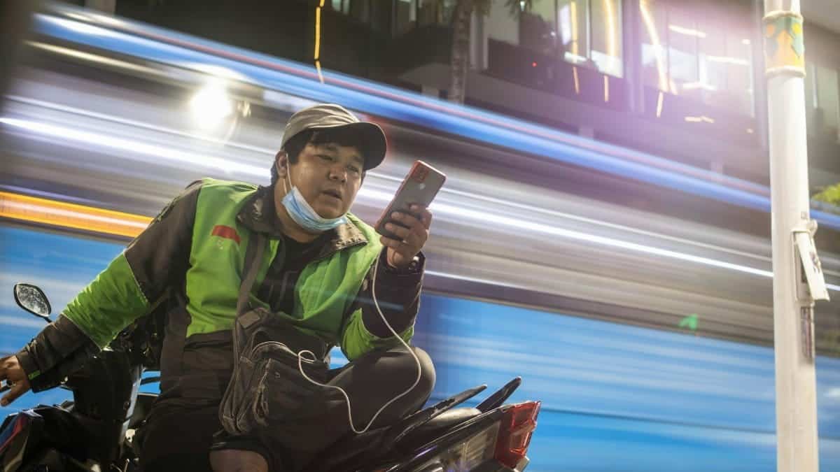 A motorcyclist checks his phone as a bus passes, creating light trails at night.
