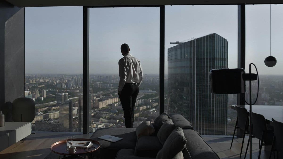 Man in a modern apartment with a panoramic view of an urban skyline and skyscraper.