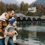 Father holds child by a scenic lake with autumn foliage, enjoying outdoor family time.