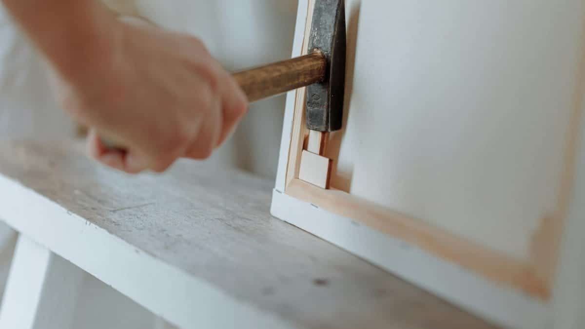 Detailed image of a hand using a hammer to work on a canvas frame in a workshop setting.