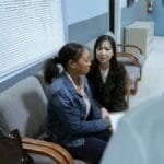 Two women in a clinic waiting room speaking with a doctor, showcasing healthcare interaction.