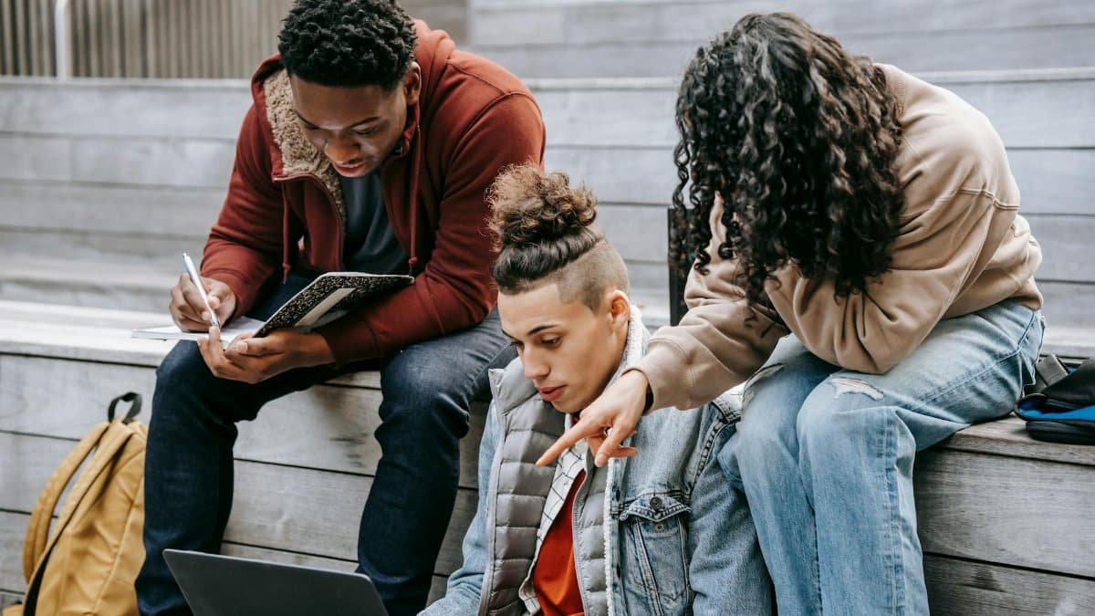 Serious multiracial group of students surfing laptop and taking notes in notebook while sitting on wooden steps during studies on street