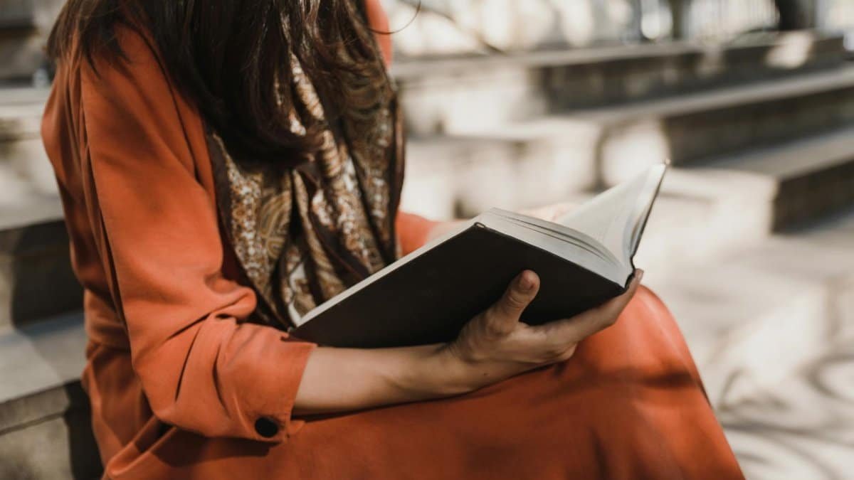 A woman wearing an orange dress is reading a book on outdoor stone steps in the sunlight.
