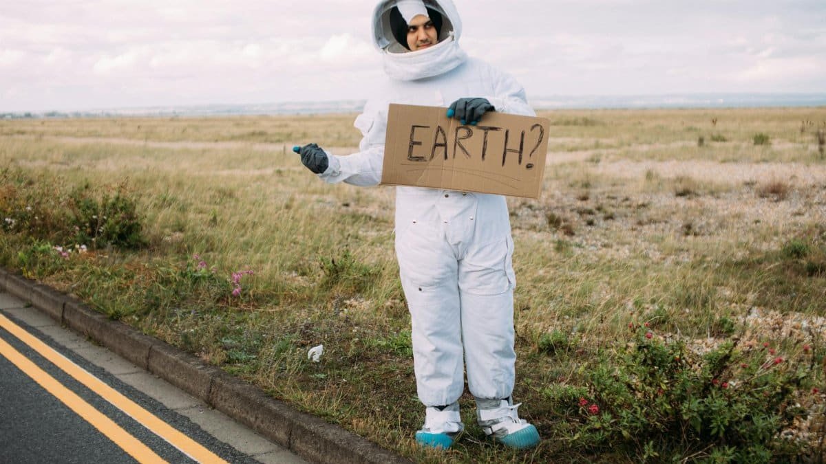 Astronaut in spacesuit hitchhiking on a rural road holding a cardboard Earth sign, symbolizing travel and adventure.