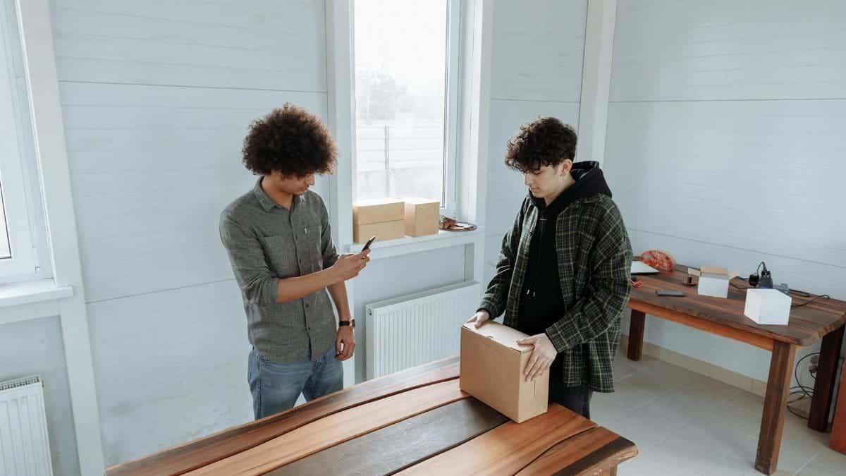 Two men unpacking boxes in a bright room, one using a mobile phone. Ideal for lifestyle or technology themes.
