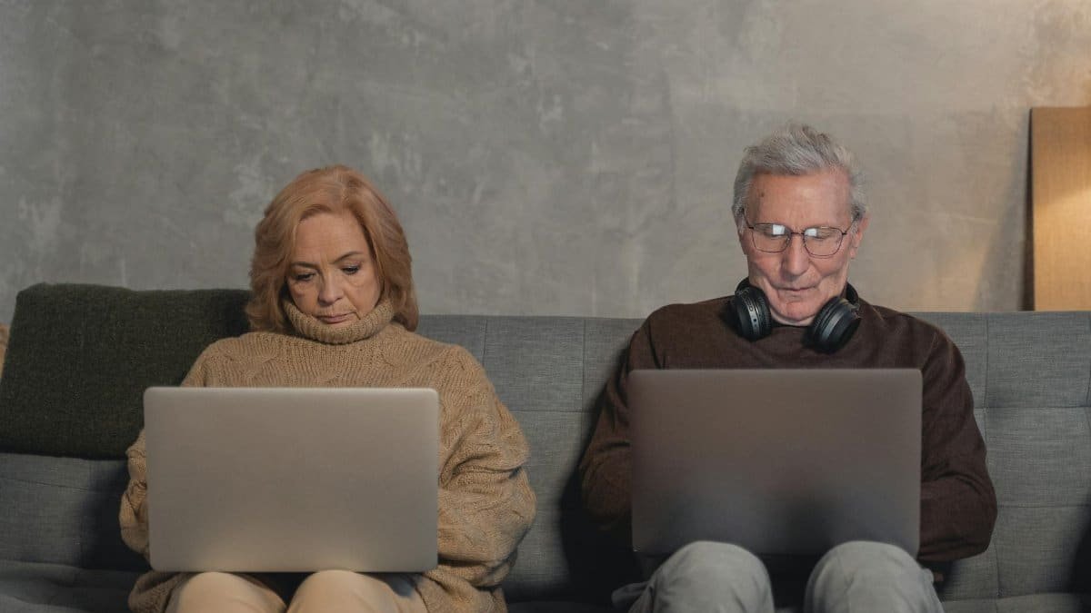 An elderly couple sitting on a sofa at home, each using a laptop. Modern technology engagement.