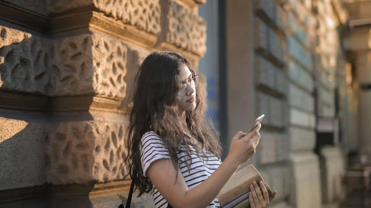 Casual portrait of a young woman leaning on a wall, using her smartphone and holding books outdoors.