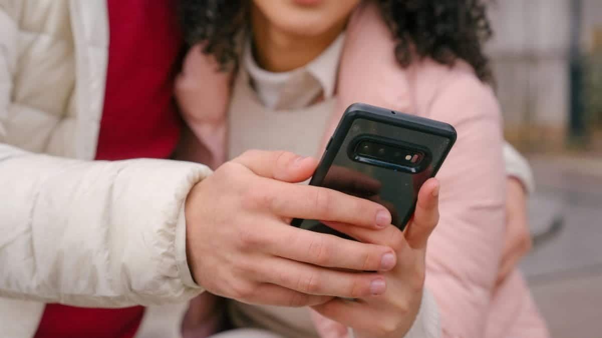 Crop anonymous young ethnic couple in casual clothes hugging and watching video on smartphone while spending time in city park