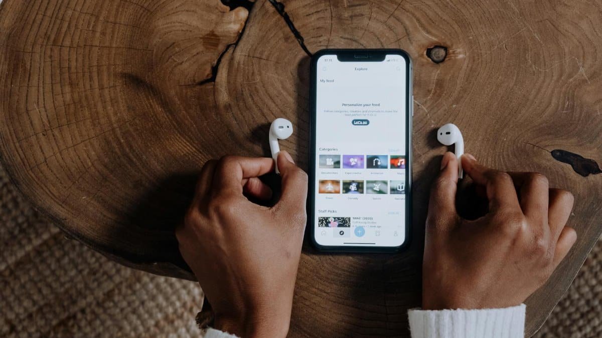 Hands holding wireless AirPods next to a smartphone displaying a social app on a wooden surface.