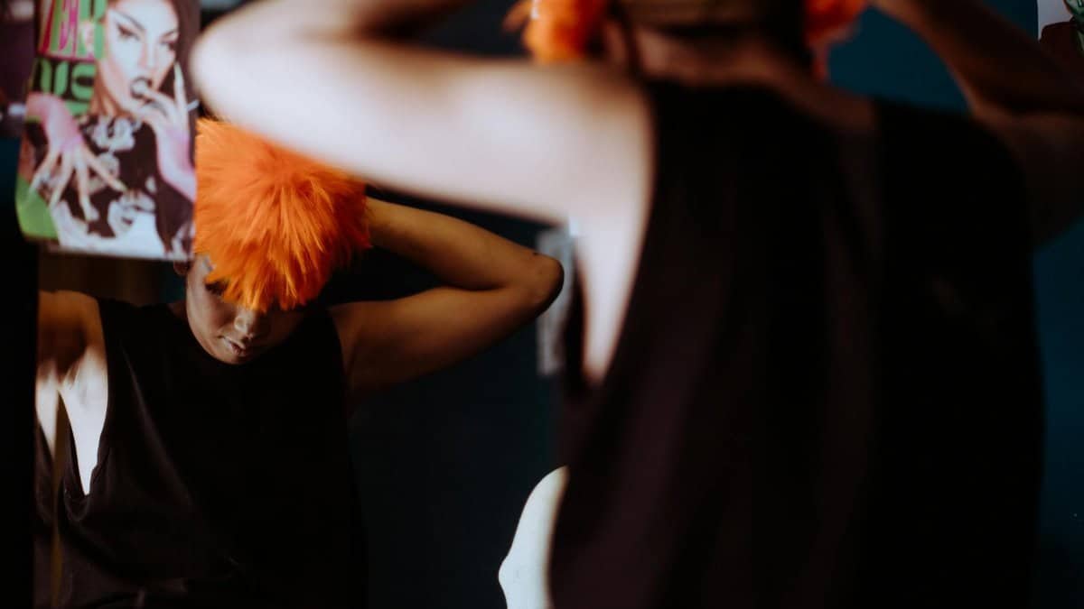 A drag performer in front of a mirror adjusting an orange wig, embodying style and individuality.
