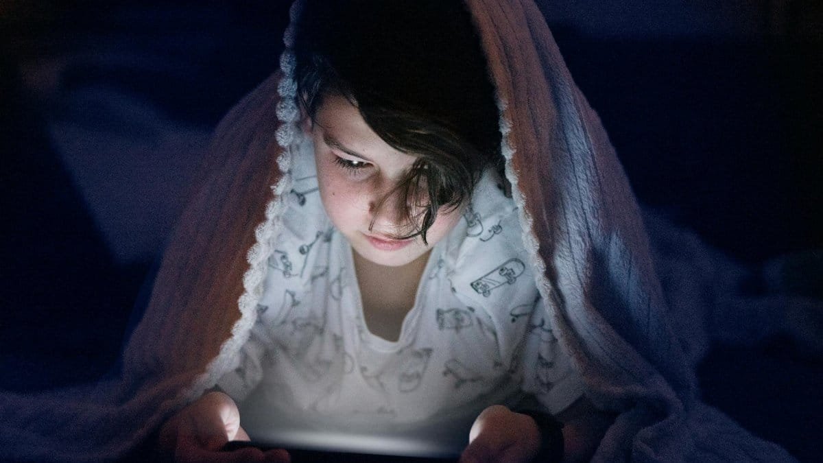 Young child under a blanket playing on a smartphone in the dark. Nighttime tech use.