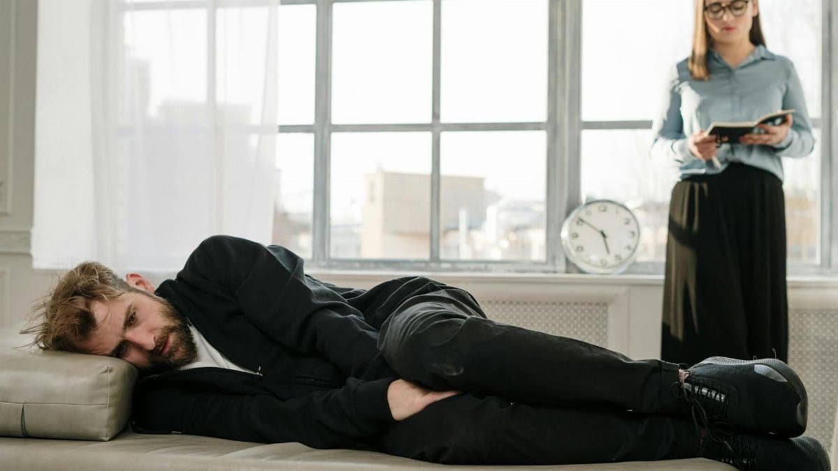 Psychological therapy session with a male patient lying on a couch and a female therapist taking notes.