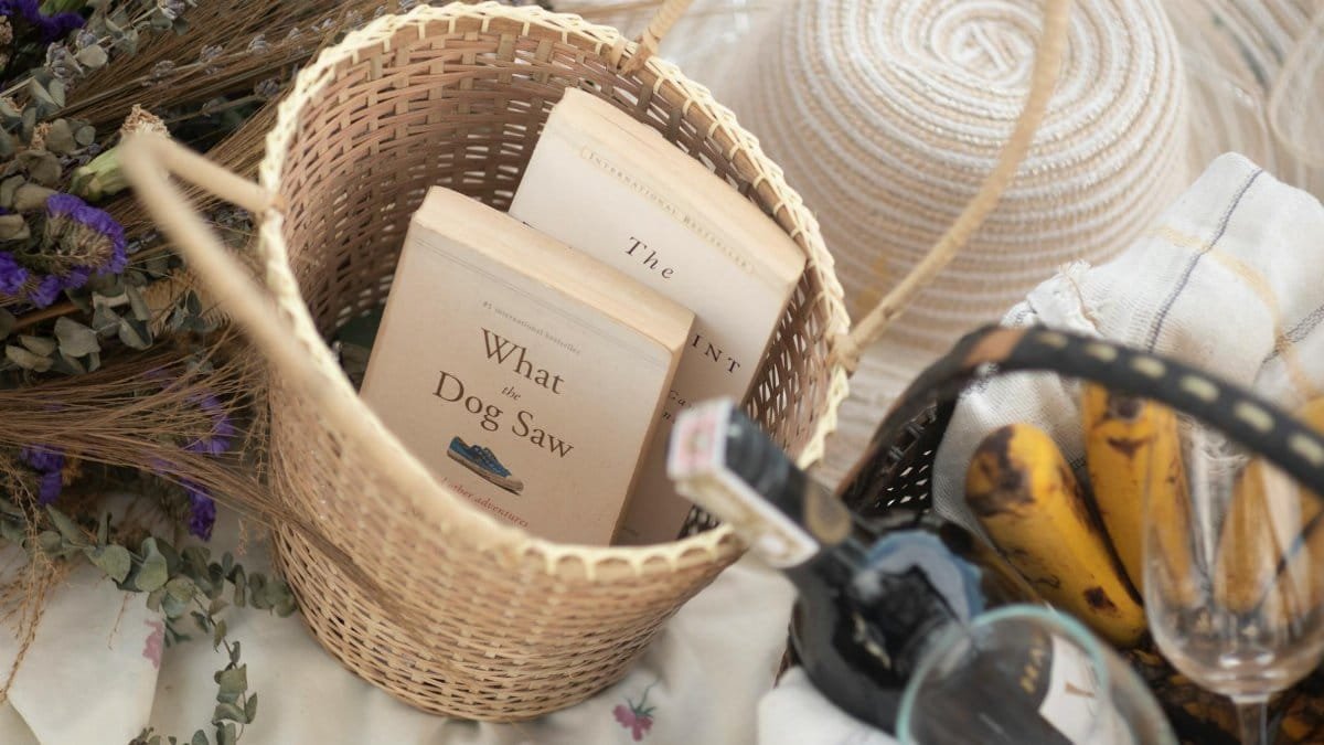 Top view of a wicker basket with books, bananas, wine, and glasses, perfect for a relaxed picnic.