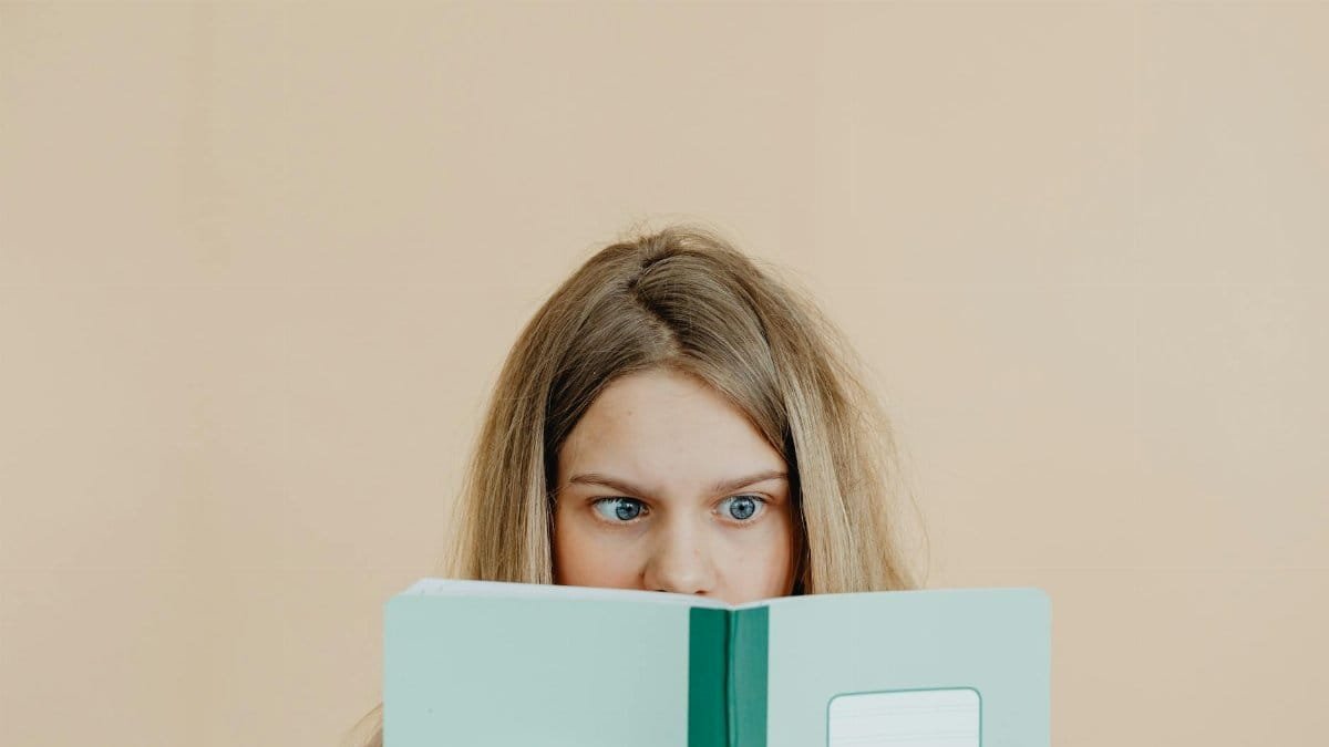Teen girl with blonde hair reading a green notebook, focused and curious.
